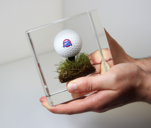 Clear acrylic commemorative gifts with golf ball, golf tee and turf embedded being held by 2 hands