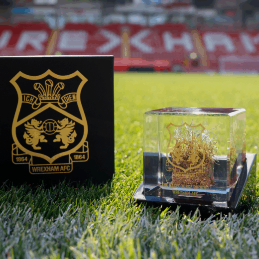 Limited edition Wrexham AFC turf cube with gold crest engraving, encasing authentic stadium turf, presented in sunlight on the Racecourse Ground.