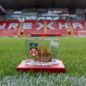 Close-up of a Wrexham AFC turf memorabilia cube with club crest, containing preserved turf from The Racecourse Ground, sitting on the pitch.