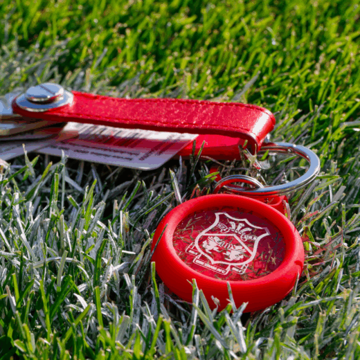 Red acrylic Wrexham AFC keyring featuring embedded turf and crest, photographed on the stadium grass as part of the fan gifting collection.