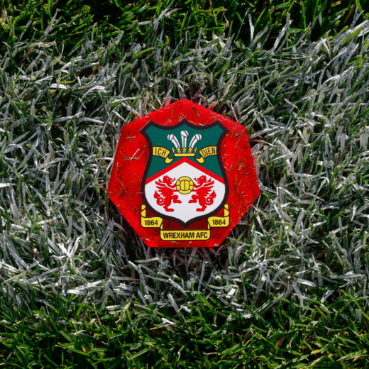 A red acrylic fan medal featuring the Wrexham AFC crest, set on the pitch turf, representing collectible football memorabilia for supporters.